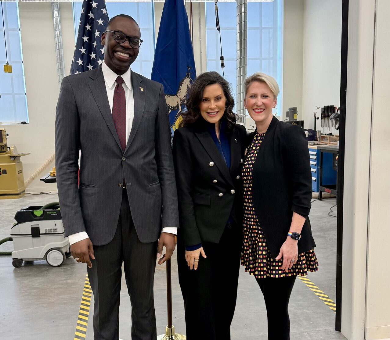 Christina Fair with Governor Gretchen Whitmer and Lt. Governor Gilchrist at a bill signing event in an industrial setting with flags behind them.