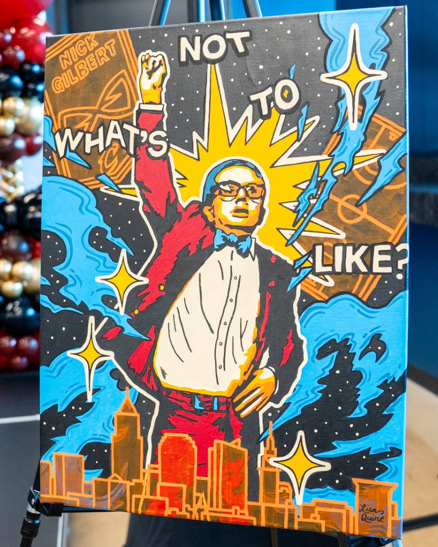 A colorful, pop-art style portrait of Nick Gilbert wearing a red blazer, blue bow tie, and glasses, with his fist raised confidently in the air. Behind him, bright yellow starbursts and comic-style elements radiate energy, along with the words “WHAT’S NOT TO LIKE?” in bold white letters. The Cleveland skyline appears at the bottom of the painting, and artist Lisa Quine’s signature is visible in the lower right corner.