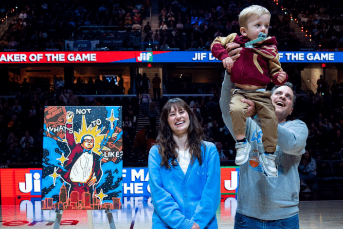 Artist Lisa Quine stands smiling at center court in Rocket Mortgage FieldHouse beside her portrait of Nick Gilbert on an easel. Her husband joyfully lifts their young son, who is wearing a Cavaliers hoodie and bow tie, into the air. The arena is filled with fans in the background, and the painting is prominently displayed beside them.