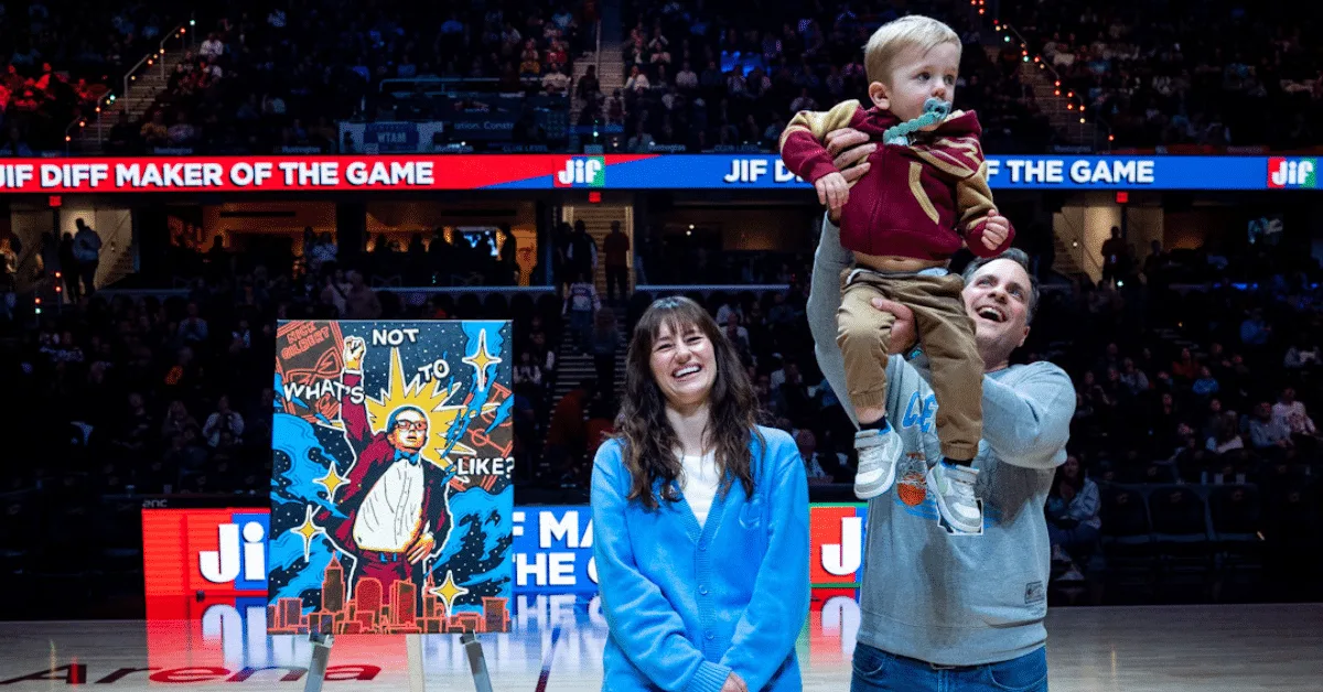 Artist Lisa Quine stands smiling at center court in Rocket Mortgage FieldHouse beside her portrait of Nick Gilbert on an easel. Her husband joyfully lifts their young son, who is wearing a Cavaliers hoodie and bow tie, into the air. The arena is filled with fans in the background, and the painting is prominently displayed beside them.
